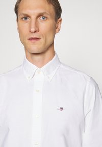 Homme à la peau claire et aux cheveux bruns courts portant une chemise blanche boutonnée avec un petit logo brodé sur la poitrine.