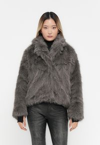 Kort grå faux fur jakke med bred krave, struktureret overflade og lange ærmer, båret over en sort turtleneck og jeans.