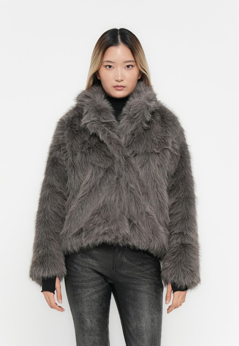 Kort grå faux fur jakke med bred krave, struktureret overflade og lange ærmer, båret over en sort turtleneck og jeans.