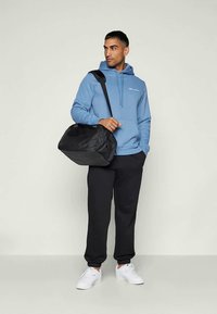 Blå huvtröja med logotyp, svart träningsväska, svarta sweatpants och vita sneakers. Väskan har en enkel design med dragkedja.