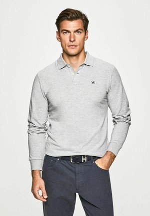 Grå långärmad polo med krage och en liten svart logotyp. Bärs med mörka byxor, vilket visar en avslappnad passform och mjuk textur.