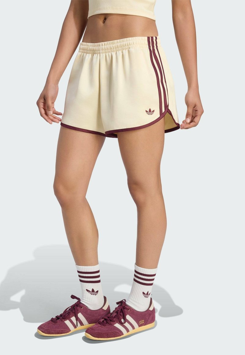 Femme portant un short crème avec des bordures et des rayures bordeaux, des baskets assorties bordeaux et crème, et des chaussettes blanches avec des rayures bordeaux et un logo.