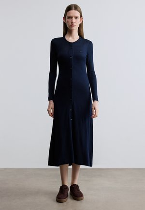 Robe à manches longues bleu marine en tissu texturé, présentant un devant boutonné, une coupe ajustée et une longueur midi. Associée à des baskets bordeaux.