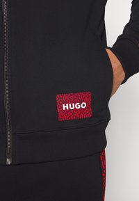 Sudadera negra con cremallera y tejido texturizado. El bolsillo izquierdo cuenta con un parche de logo en rojo y negro, con un patrón llamativo que muestra "HUGO" en blanco.