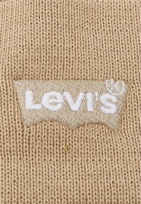 Tessuto beige con lavorazione a maglia testurizzata, caratterizzato da un logo Levi's ricamato in bianco su uno sfondo beige a contrasto, in una forma curva simile a un distintivo.