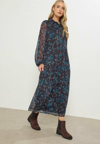 Maxi abito floreale scuro con lunghe maniche a palloncino, motivi blu e verde petrolio, e un design a strati, abbinato a stivali alla caviglia marroni.