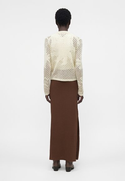 Γυναίκα στέκεται με την πλάτη, φοράει ένα κρεμ πλεκτό μακρυμάνικο τοπ και μια καφέ μάξι φούστα με ψηλή μέση και πλαϊνή σχίσιμο, μαύρα παπούτσια με τακούνια.