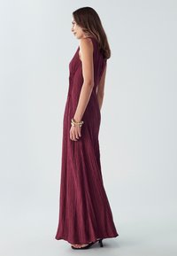 Dlhé, bezrukávové burgundské šaty s textúrovaným zmršteným dizajnom, priliehavým korzetom a voľnou sukňou, doplnené o čierne sandále na podpätku.