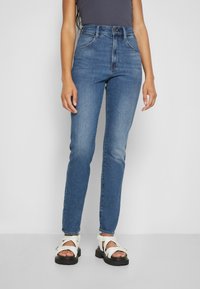 Høytlivte blå denimjeans med slim fit, med kontrastsøm og fem-lomme-design, kombinert med hvite chunky sandaler.
