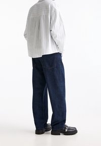 Personne portant un jean bleu foncé ample, des mocassins noirs épais, et une chemise blanche à manches longues avec de fines rayures verticales, se tenant les mains dans les poches.