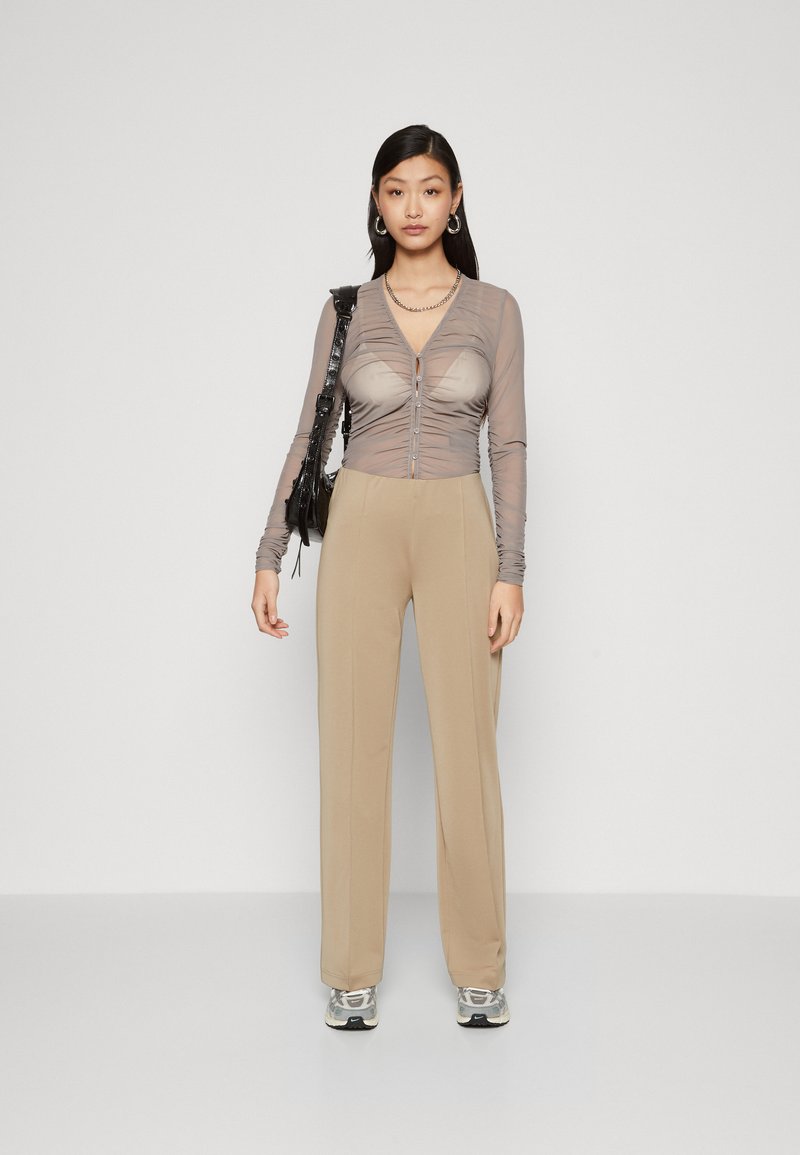 Durchsichtige, leichte taupefarbene Bluse mit langen Ärmeln, Knopfverschluss und Raffung. Beigefarbene, hochtaillierte Hose und graue Sneaker. Schwarze Tasche mit Trageriemen.