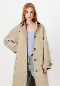 Beige trenchcoat met groene bloemborduursels, grote knopen en een losse pasvorm. Beschikt over een brede kraag en iets oversized mouwen.