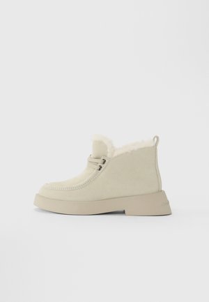 Beige suède enkelboot met dikke zool, bontvoering rond de enkel, vetersluiting aan de voorkant en een trekkinglus aan de hiel.