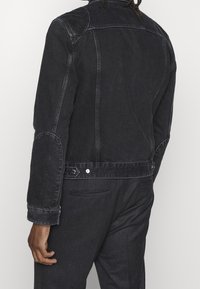 Svart denimjacka med en figurnära passform, böjda axelsömmar, armbågsfleece och knappr i midjan. Texturerad tyg, minimal sömnad.