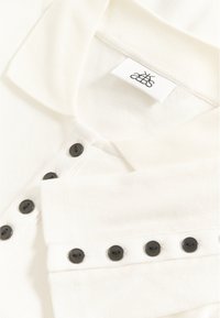 Biała żebrowana koszulka polo z długim rękawem, z czarnymi guzikami z przodu i na mankietach, zbliżenie na kołnierzyk i metkę.