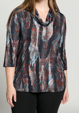Vrouw die een top met lange mouwen draagt met zwarte, grijze en rode paisley patronen en een zwarte broek tegen een effen lichte achtergrond.