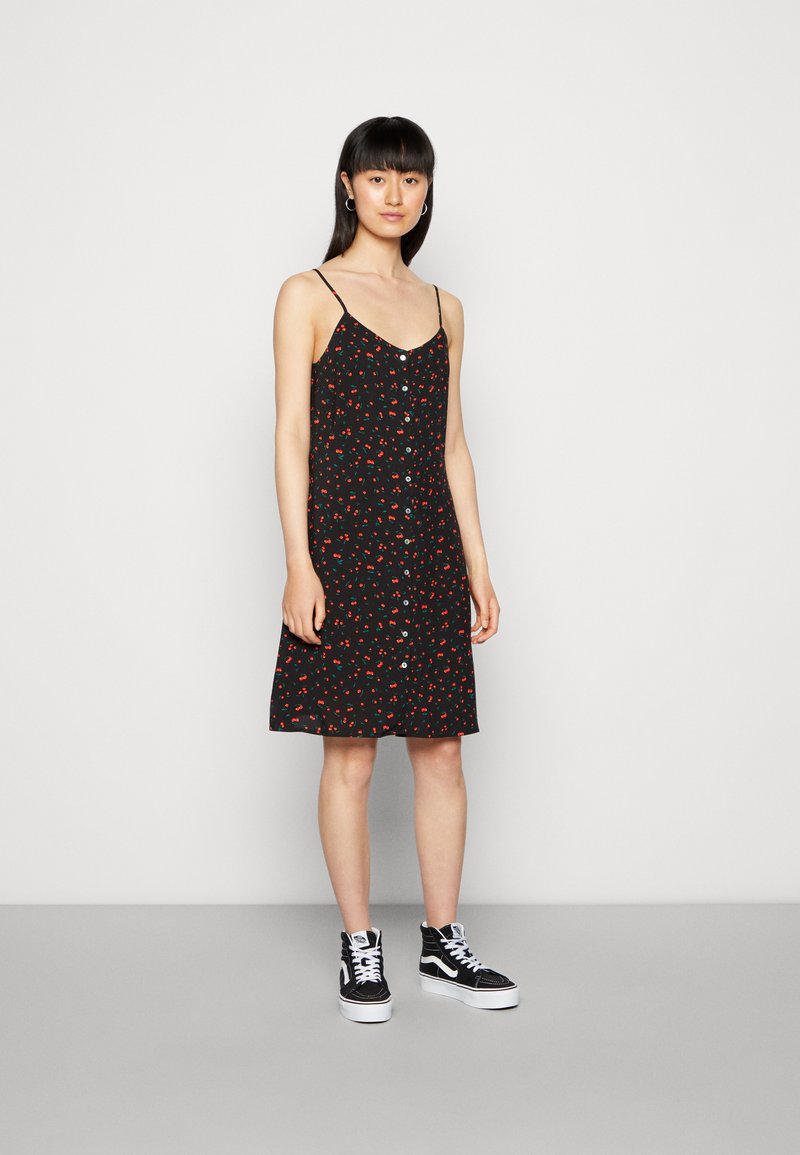 Vestido preto sem mangas com um pequeno padrão de flores vermelhas, detalhe de botões na parte da frente e uma silhueta em A. Combinado com ténis pretos de cano alto.