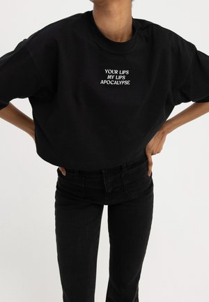 Sort croppet t-shirt med rund halsudskæring, prydet med hvidt broderet tekst: "YOUR LIPS MY LIPS APOCALYPSE." Kombineret med sorte jeans.