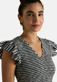 V-Ausschnitt-Kleid mit schwarz-weißem Karomuster, Rüschensäumen, strukturierter Stoffqualität und gerafftem Detail an der Taille.