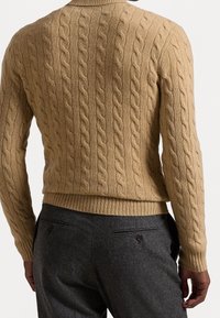 Beżowy sweter w splot warkoczowy z żeberkowym dekoltem i dołem, prezentowany od tyłu, w zestawieniu z szarymi spodniami z zapinanymi kieszeniami z tyłu.
