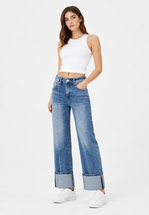 Kvinde stående med hånd i lommen iført hvid ærmeløs crop top, brede blå jeans med opsmøg og hvide sneakers på ensfarvet baggrund.