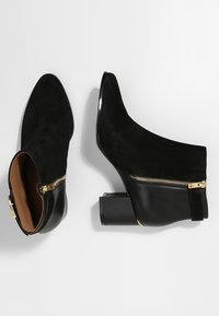 Botas negras hasta el tobillo con punta afilada, parte superior de ante, detalle de cremallera dorada y tacón ancho. Textura suave con un ribete de cuero contrastante.