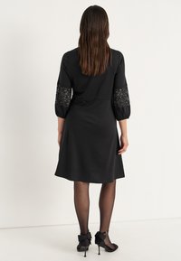 Musta mekko, jossa on 3/4 pitsihihat, kellotettu helma ja sileä rakenne. Yhdistettynä mustiin sukkahousuihin ja korkokenkiin, joissa on suuret nauha-akkentit.