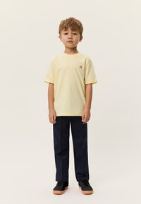 Ανοιχτό κίτρινο βαμβακερό μπλουζάκι με ρεβέρ στα μανίκια και μικρό κόκκινο λογότυπο. Φορεμένο με ναυτικά μπλε παντελόνια και μαύρα αθλητικά παπούτσια με λαστιχένιες σόλες.