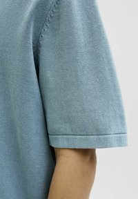 Un pull tricoté à manches courtes de couleur bleu clair qui présente une texture lisse, avec des bords de manches roulés et de subtils détails de couture le long de l'épaule.