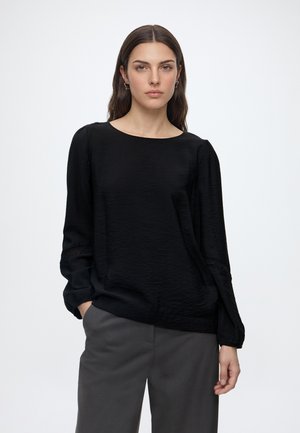 Femme aux longs cheveux foncés portant un chemisier noir à manches longues et un pantalon gris, debout avec une main dans la poche sur fond blanc.