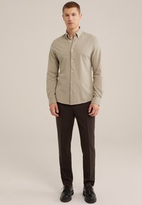 Cămașă bej, cu guler, din material neted, cu mâneci lungi, croi slim, cu un logo brodat subtil, asociată cu pantaloni maron închis și pantofi negri.