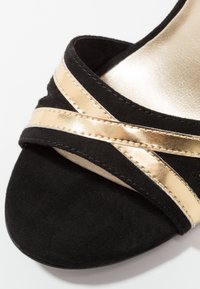 Sandale noire avec des brides métalliques dorées croisées et une semelle intérieure rembourrée visible en gros plan.