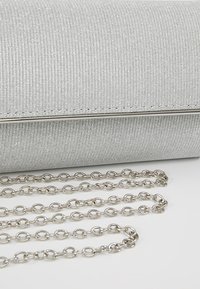 Pochette texturée argentée avec bordure en métal et longue bandoulière chaîne argentée étalée sur une surface blanche.