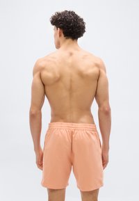 Άνδρας με σγουρά μαλλιά, γυμνό πάνω μέρ ος του σώματος, φοράει μαγιό σε ροζ χρώμα, στέκεται με την πλάτη προς τα εμπρός σε ένα απλό ανοιχτό φόντο.