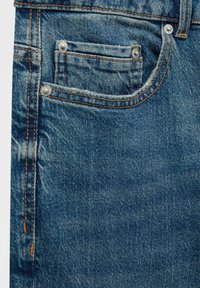 Gros plan sur une poche de jean en denim bleu avec des coutures visibles, des rivets et une petite poche à monnaie à l'intérieur de la poche avant droite.