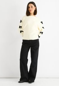 Cream sweater with black bows on sleeves, ribbed hem, and loose fit, paired with high-waisted black wide-leg pants and black flats.