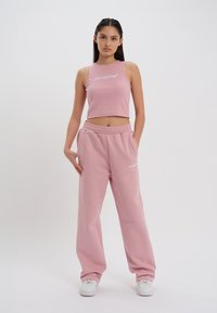 Top scurt roz și pantaloni dintr-un material moale, cu picioare largi, asortați, având un logo pe top și pe pantaloni. Tenisuri albe completează aspectul.