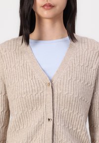 Beige knitted cardigan with a textured pattern, featuring a deep V-neck and two gold heart-shaped buttons at the front.