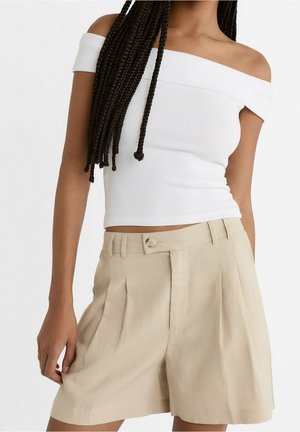 Woman wearing a white off-shoulder top and beige high-waisted pleated shorts with button closure, standing with one hand by her side.