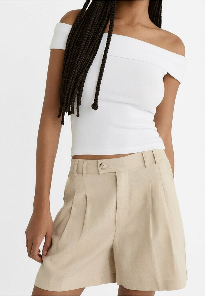 Femme portant un haut blanc épaules dénudées et un short plissé beige taille haute avec fermeture à boutons, debout avec une main le long du corps.