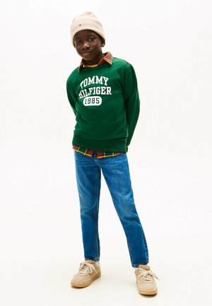 Zielona bluza z kapturem z napisem "TOMMY HILFIGER 1985" w kolorze białym, założona na koszulę w kratę z flaneli. Noszona z niebieskimi dżinsami i beżowymi sneakersami.