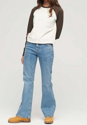 Femme portant un jean évasé bleu, un t-shirt raglan blanc et marron foncé à manches longues, et des bottes de travail marron clair, debout devant un fond uni.