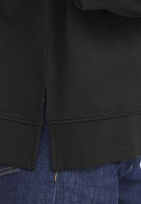 Felpa nera con taglio dritto, fessure laterali e texture liscia, abbinata a jeans in denim blu con un evidente dettaglio di cucitura gialla.