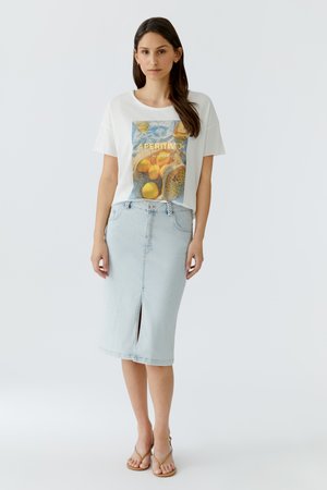 Femme aux longs cheveux foncés portant un t-shirt blanc graphique avec des fruits et du texte, une jupe midi en jean clair avec une fente devant, et des sandales beiges.