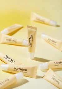 Torriden Solid In Lip Essence i en lysegul tube med et hvidt fliplåg og trykt mærkning, arrangeret på en blød lysegul baggrund.