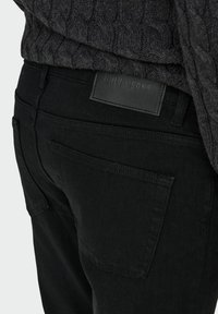 Pantalones de mezclilla negros con primer plano del bolsillo trasero y la etiqueta de la marca. La tela tiene un acabado suave, con un diseño clásico de 5 bolsillos.