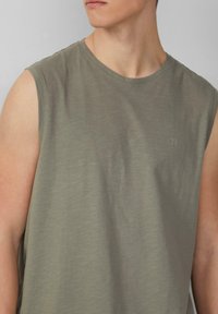 Olijfgroene mouwloze t-shirt gemaakt van zachte stof; heeft een ronde halslijn en een klein "QS" logo op de borst. Soepele textuur.