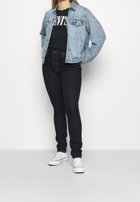 Džínová bunda světle modré barvy, černé tričko s potiskem, černé skinny džíny s vysokým pasem a bílé tenisky; pohodlný vrstvený outfit.