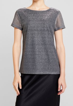 Mujer con una blusa gris brillante de manga corta y cuello redondo, combinada con una falda negra, de pie frente a un fondo gris.