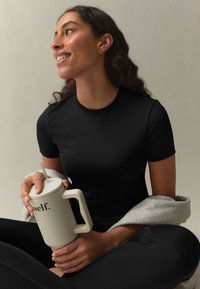 Svart figurnära kortärmad topp, kombinerad med svarta leggings. Modellen håller en stor beige mugg med texten "self." tryckt i svart, sittande på en ljus bakgrund.